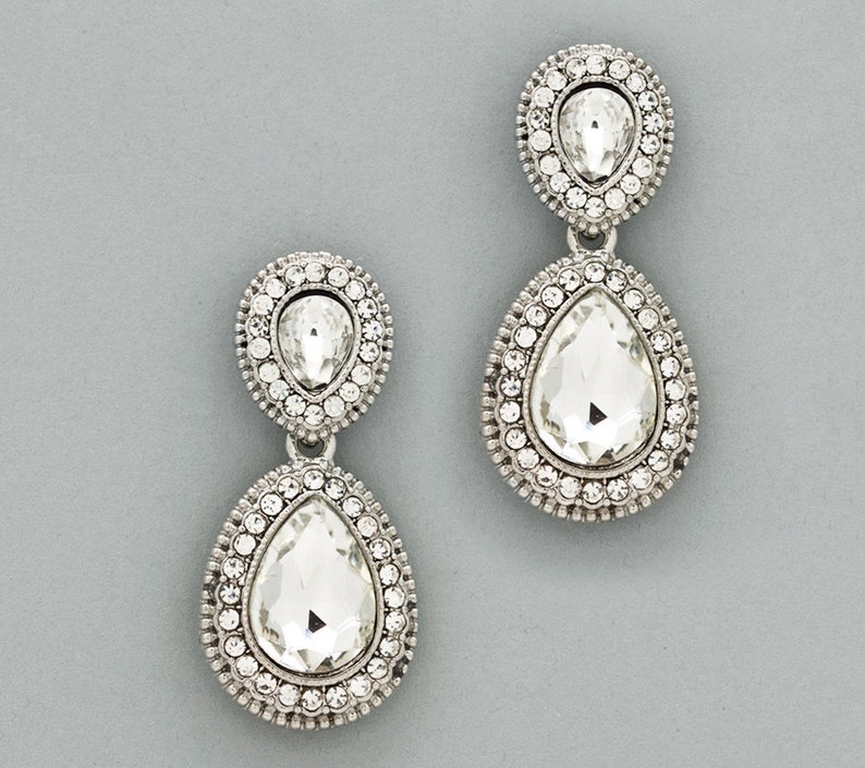 May include: A pair of silver teardrop earrings with clear crystal accents. Each earring features two teardrop-shaped crystals, one smaller than the other, surrounded by a halo of smaller crystals.