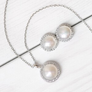 Puede incluir: Un collar y pendientes de plata con una perla grande de color blanco rodeada por un halo de peque&ntilde;as piedras transparentes.