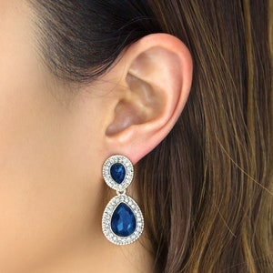 May include: A pair of dangle earrings with two teardrop-shaped blue crystals surrounded by smaller clear crystals. The earrings are gold-toned.