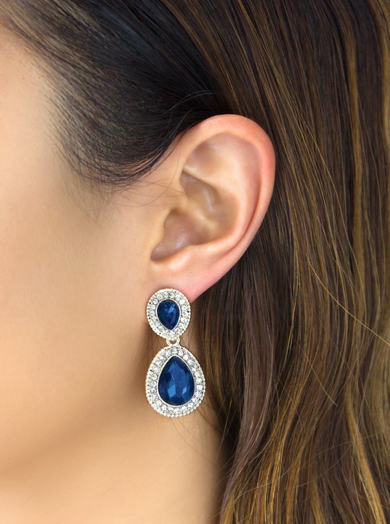 May include: A pair of dangle earrings with two teardrop-shaped blue crystals surrounded by smaller clear crystals. The earrings are gold-toned.