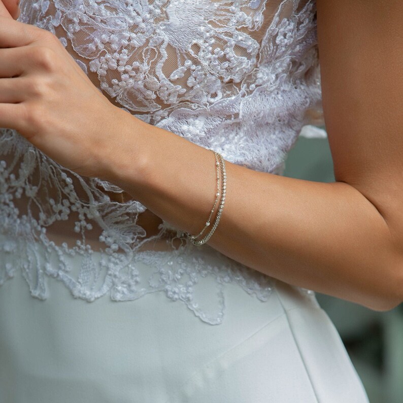 Peut inclure: Un bracelet d&eacute;licat &agrave; deux rangs avec de petites pierres scintillantes. Le bracelet est port&eacute; sur un bras, sur fond de robe en dentelle blanche. Les pierres semblent &ecirc;tre claires, refl&eacute;tant la lumi&egrave;re.