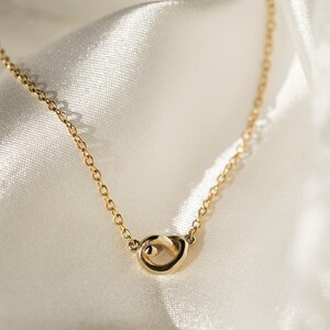 May include: A delicate gold chain necklace with a simple, abstract knot pendant. The necklace is displayed on a white satin background.