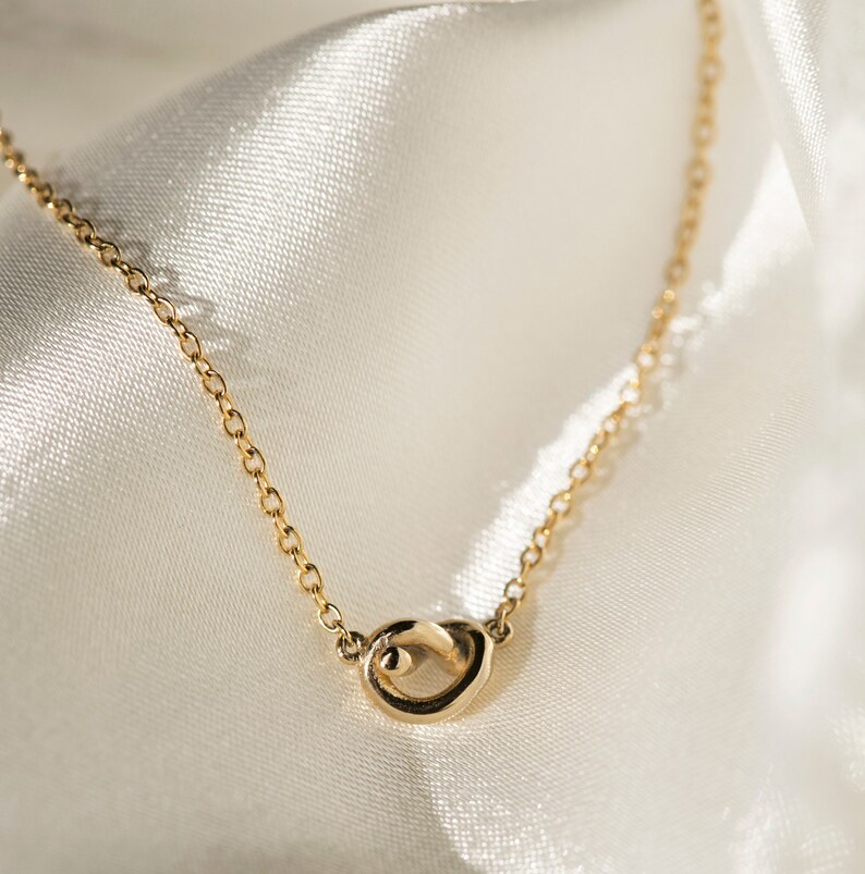May include: A delicate gold chain necklace with a simple, abstract knot pendant. The necklace is displayed on a white satin background.