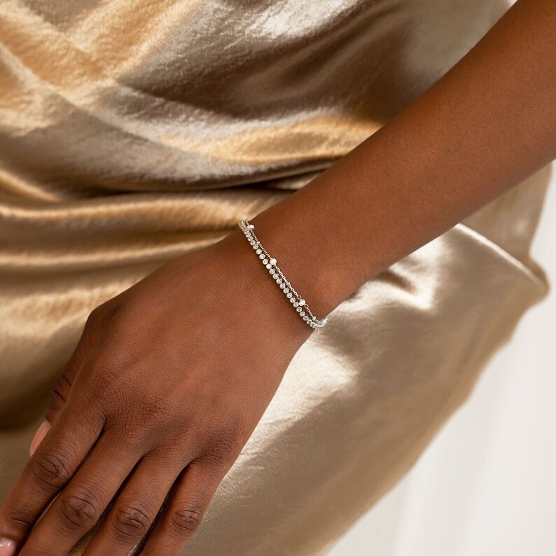 Peut inclure: Un bracelet en argent avec deux rang&eacute;es de petites pierres scintillantes. Le bracelet est port&eacute; au poignet d'une personne, sur un fond de tissu soyeux de couleur or. Le bijou est d&eacute;licat et &eacute;l&eacute;gant.