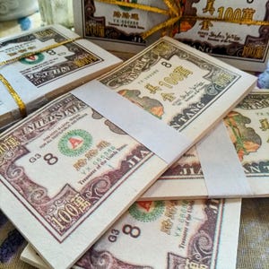 May include: Stacks of novelty $100 bills, bound with white paper bands and gold ribbon. The bills feature Chinese characters and the words "The United States of America." The bills are tan with brown and green accents.
