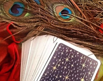 May include: A deck of tarot cards with a dark blue background and gold starburst design. The deck is fanned out, with peacock feathers in the background. The feathers have brown, blue, and green accents. A red fabric is visible.