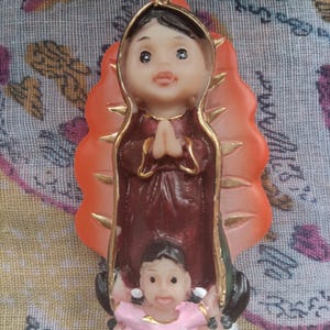 May include: A religious figurine of Our Lady of Guadalupe, featuring a detailed depiction of the Virgin Mary in a maroon robe with gold accents. The figurine includes a smaller figure at the base and the name "Guadalupe".