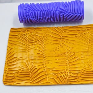 Könnte beinhalten: Eine lila strukturierte Walze und ein rechteckiges Stück gelbes Material mit einem Blattmuster. Die Walze ist zylindrisch mit einem sich wiederholenden Blattdesign. Das gelbe Material hat ein erhabenes Blattmuster.