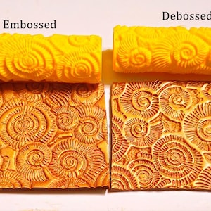 May include: Two yellow textured rolling pins, labelled "Embossed" and "Debossed", are above two rectangular clay impressions. The clay impressions are orange and feature a spiral design.