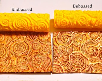 May include: Two yellow textured rolling pins labeled "Embossed" and "Debossed" are displayed above two rectangular clay impressions. The clay impressions are orange and feature a spiral design.