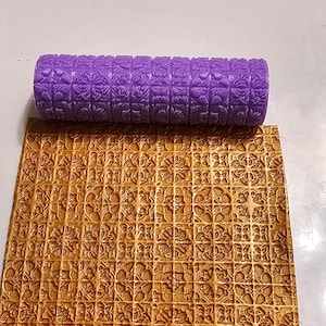 May include: Purple silicone rolling pin with a repeating geometric pattern. The rolling pin is used to create an embossed design on fondant or dough. The image also shows a piece of fondant with the embossed design.
