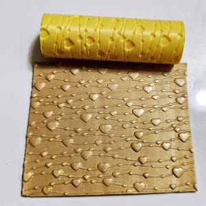 Peut inclure: Un rouleau à pâtisserie jaune avec un motif de cœur et de chaîne. Le rouleau à pâtisserie est utilisé pour créer un motif en relief sur une feuille de fondant dorée.