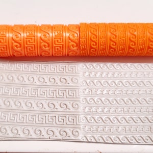May include: Orange rolling pin with a repeating pattern of Greek key and wave designs. The rolling pin is used to create an embossed pattern on a white surface.