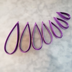 Könnte beinhalten: Ein Satz mit acht violetten Plastik-Ausstechern in Tropfenform, die in einem Halbkreis angeordnet sind.