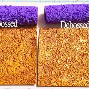 Może przedstawiać: Dwa fioletowe wałki z wzorem, oznaczone jako "Embossed" i "Debossed", znajdują się nad dwoma prostokątnymi odciskami gliny. Odbitki gliniane są w kolorze złotym i mają szczegółowy wzór kwiatowy i wirujący. Tekst "Embossed" i "Debossed" jest biały.