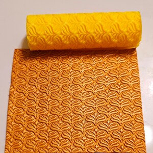 May include: A yellow rubber roller with a repeating leaf pattern, used for creating an embossed design on a gold surface. The surface has a repeating leaf pattern.