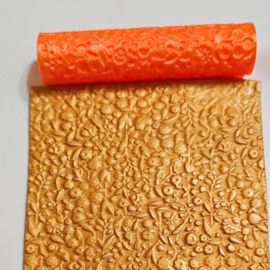 Puede incluir: Rodillo naranja con un diseño floral, utilizado para crear un patrón en relieve en una superficie dorada.
