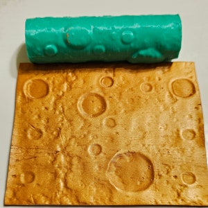 Puede incluir: Un rodillo verde azulado con una superficie texturizada, utilizado para crear un patrón lunar en una superficie dorada. La superficie tiene un aspecto texturizado y con cráteres.
