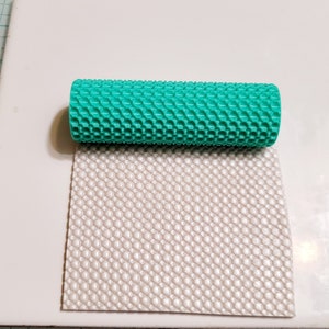 Puede incluir: Un rodillo turquesa con un patrón de panal, junto a una hoja de fondant blanco texturizado.