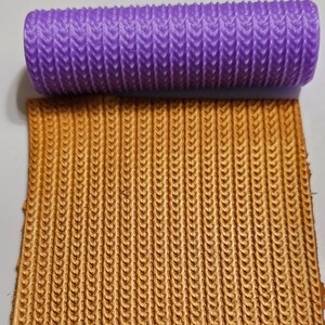 May include: A purple silicone rolling pin with a textured chevron pattern. The rolling pin is used to create a textured pattern on a gold surface.