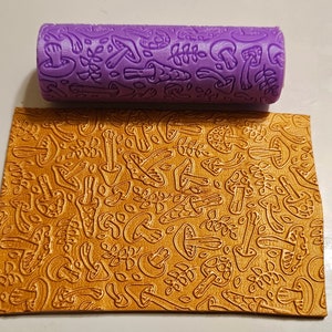 Pode incluir: Um rolo de pastelaria roxo com um desenho de cogumelos e folhas. O rolo é usado para criar um padrão em relevo numa folha de fondant dourada.