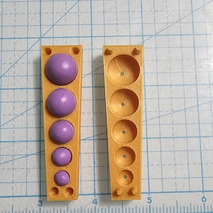 May include: Two yellow plastic molds with five round depressions. The mold on the left has five purple spheres in the depressions. The molds are on a gridded surface.