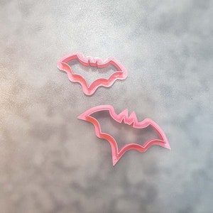 May include: Two pink plastic bat-shaped cookie cutters. The cutters are on a gray surface.