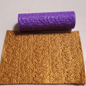 May include: A purple rolling pin with a floral pattern sits above a sheet of gold-colored material with a matching embossed floral design. The rolling pin is cylindrical and features a repeating pattern of flowers and leaves.