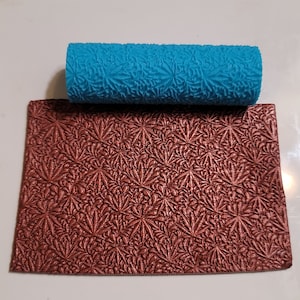 Puede incluir: Un rodillo azul con un patrón floral y una hoja texturizada marrón a juego. El rodillo se utiliza para crear patrones decorativos en fondant o arcilla.
