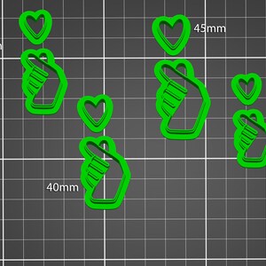 May include: Green plastic cookie cutters in the shape of a hand making a heart symbol and individual hearts. The hand shapes are 40mm and 45mm wide. The hearts are 30mm and 35mm wide. The background is a gray grid.