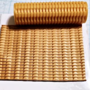 May include: A tan-coloured roller with vertical ridges rests above a sheet of material with a matching embossed pattern. The sheet has a repeating square pattern, creating a textured surface. The roller and sheet are both in a tan colour.