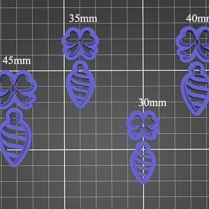 Op de afbeelding: Een verzameling paarse kerstkoekjessnijders in de vorm van ornamenten en strikken, in verschillende maten van 20 mm tot 45 mm. De snijders zijn gerangschikt op een rasterachtergrond, met hun afmetingen.