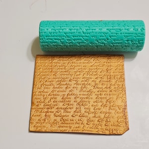 Puede incluir: Un rodillo turquesa y una pieza rectangular de madera, ambos con una textura similar a la escritura. El rodillo es cilíndrico y la madera es de color marrón claro. La escritura parece ser un patrón repetitivo.