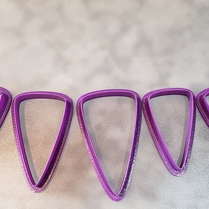 Pode incluir: Conjunto de seis cortadores de biscoitos em forma de triângulo roxo de tamanhos diferentes.