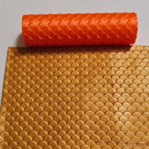 以下が含まれることがあります： オレンジ色のテクスチャローラーのクローズアップ。魚鱗模様が刻まれています。このローラーは、金色の表面に同様の模様を作成するために使用されます。