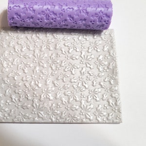 May include: A purple silicone rolling pin with a holly leaf and berry design. The rolling pin is used to create an embossed pattern on a white fondant sheet.
