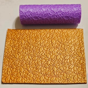 Könnte beinhalten: Eine lilafarbene Rolle mit einem Blumenmuster, die verwendet wird, um ein Prägemuster auf einer goldenen Oberfläche zu erstellen.