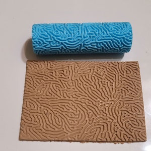 May include: A blue paint roller with a textured pattern resembling coral. The roller is shown next to a sample of the pattern created by the roller on a brown surface.