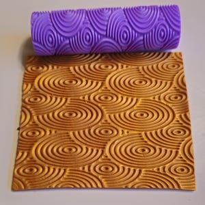 May include: A purple textured rolling pin rests above a square of embossed material. The rolling pin and the material have a concentric circle pattern. The material is a warm, golden color, and the rolling pin is a vibrant purple.