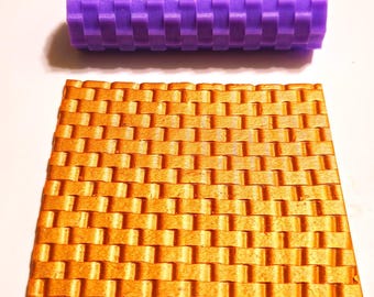 Può includere: Un mattarello strutturato viola e un foglio rettangolare color oro, entrambi con un motivo a trama di vimini. Il mattarello è cilindrico e il foglio è piatto, creando una trama visiva contrastante.