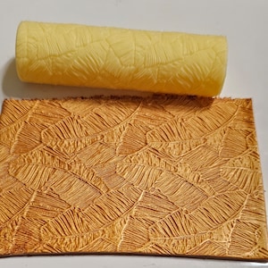 Puede incluir: Un rodillo de goma amarillo con un diseño de patrón de hojas. El rodillo se utiliza para crear un efecto texturizado en una superficie. La superficie es un material marrón similar al cuero con un patrón de hojas.