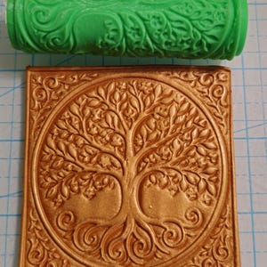 Könnte beinhalten: Eine grün gemusterte Walze und eine goldfarbene quadratische Fliese mit einem Lebensbaum-Design. Die Walze hat ein sich wiederholendes Blumenmuster. Die Fliese zeigt ein detailliertes Baumdesign in einem kreisförmigen Rahmen.