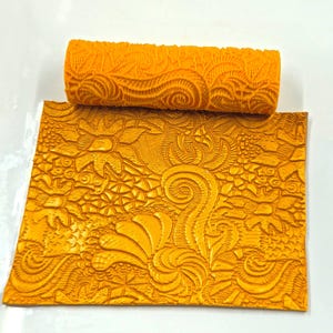 May include: A bright orange cylindrical patterned rolling pin and a matching rectangular flat sheet of material. Both items feature an intricate, embossed design of swirling lines, floral shapes, and textured patterns pressed into the surface.