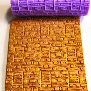 May include: A purple textured rolling pin rests above a gold-colored rectangular sheet with a matching pattern. The rolling pin and sheet feature a repeating rectangular design with what appears to be Hebrew text.