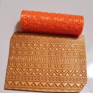 May include: Orange rolling pin with a geometric pattern, used for creating embossed designs on fondant or clay. The rolling pin is shown next to a gold-colored sheet of embossed fondant or clay.