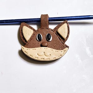 May include: A brown and beige clay cat charm with black eyes. The charm has a stitched detail around the edges and a loop at the top for hanging.
