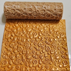 May include: A beige rolling pin with a detailed seashell pattern, alongside a sheet of dough embossed with the same design. The rolling pin and the embossed dough are in a light brown color, creating a textured effect.