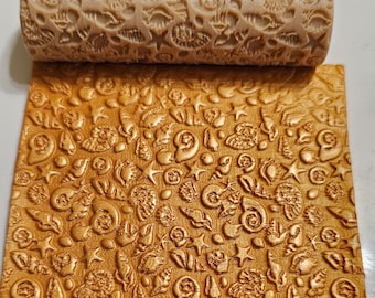 May include: A beige rolling pin with a detailed seashell pattern, alongside a sheet of dough embossed with the same design. The rolling pin and the embossed dough are in a light brown color, creating a textured effect.