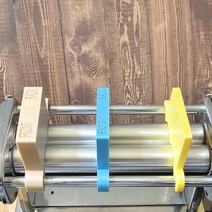 Puede incluir: Una máquina de pasta de acero con tres rodillos. Hay tres guías de colores fijadas a la máquina, una beige, una azul y una amarilla. Cada guía tiene las palabras "FRONT" y "BACK" impresas en ella.