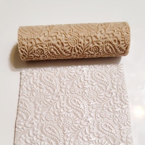 Op de afbeelding: Een bruine deegroller met een bloemmotief. De deegroller wordt gebruikt om een reliëfpatroon op fondant of deeg te creëren. De afbeelding toont ook een voorbeeld van het patroon dat door de deegroller wordt gemaakt.